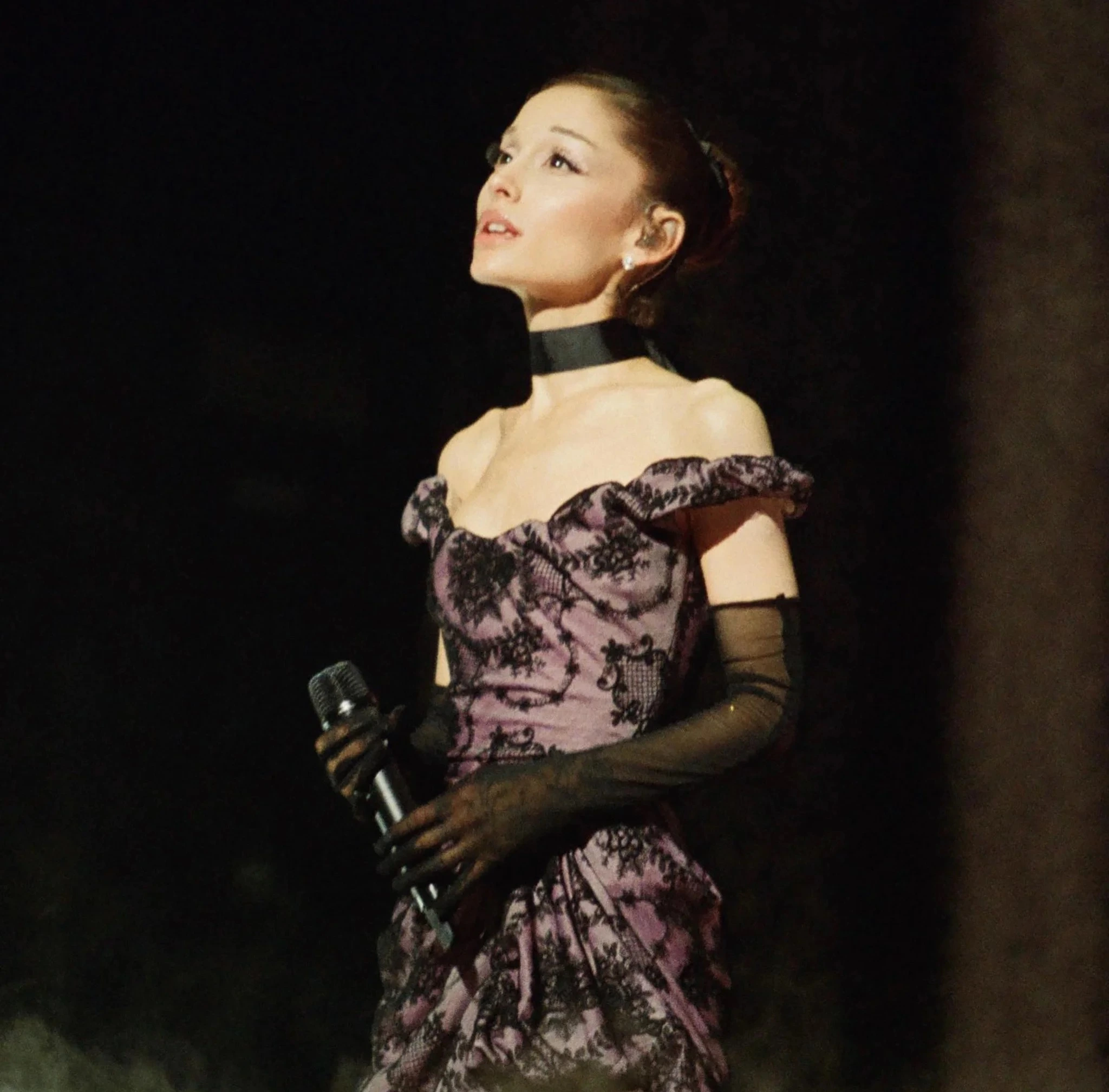 Ariana Grande performing on stage in a mauve lace dress while holding a microphone under dramatic concert lighting
